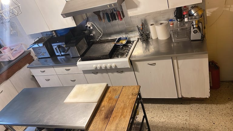 Small kitchen with white cabinets, stove, hood vent, and metal counter with wooden cutting board on a table in foreground
