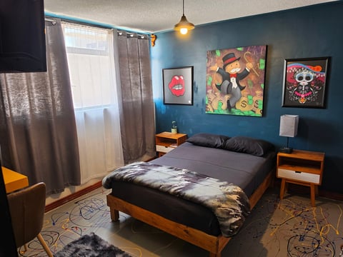 Modern bedroom with teal blue walls, wood frame bed, colorful pop art on walls, pendant light, and window with curtains