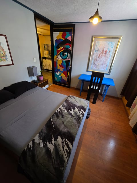 A cozy bedroom with a bed, blue desk chair, colorful door art, hardwood floor, and pendant light overhead