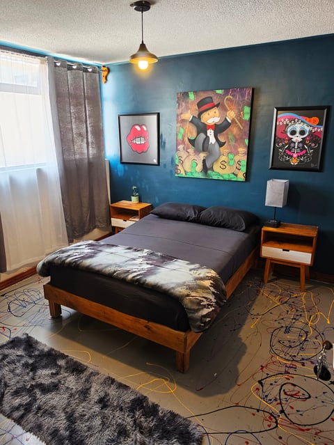 Modern bedroom with wood bed frame, dark blue walls, colorful artwork, pendant light, and patterned rug