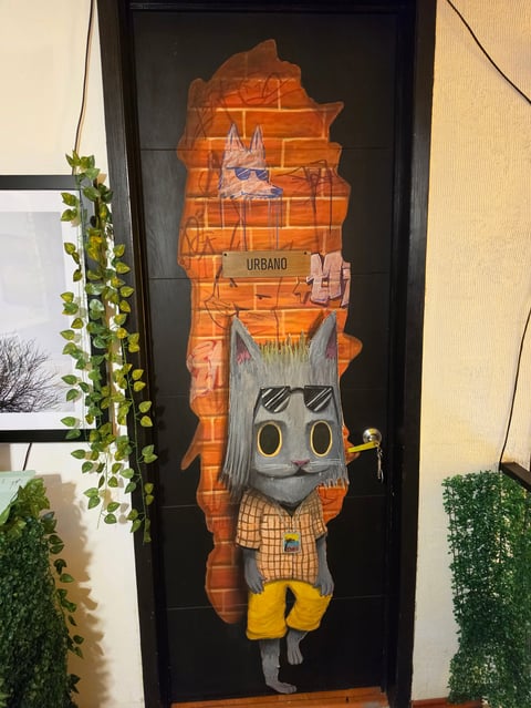 Framed street art featuring a stylized cat character with gray face, checkered clothing, and yellow boots, surrounded by an orange map silhouette with URBANO text