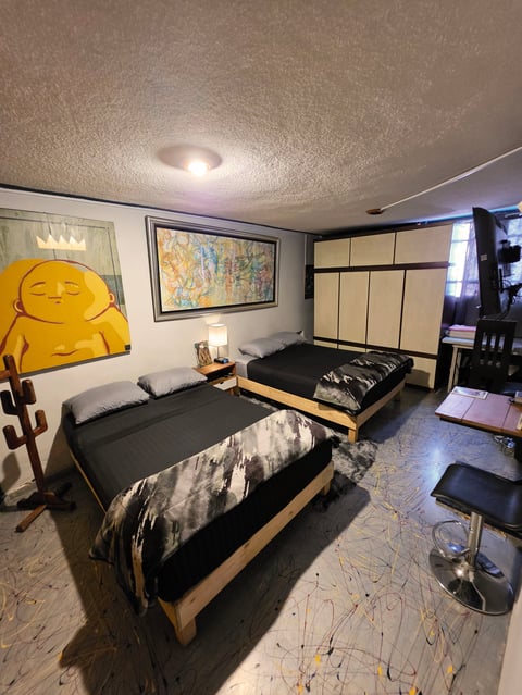 Modern bedroom with two beds, wooden frames, black bedding, yellow Buddha sculpture on wall, framed artwork, wardrobe, and ceiling light.