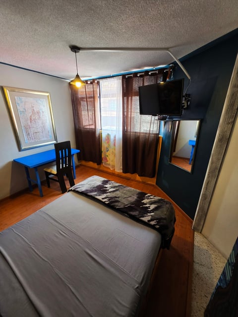 A compact bedroom with a bed, blue desk and chair, TV mounted on dark wall, and windows with brown curtains overlooking natural light.