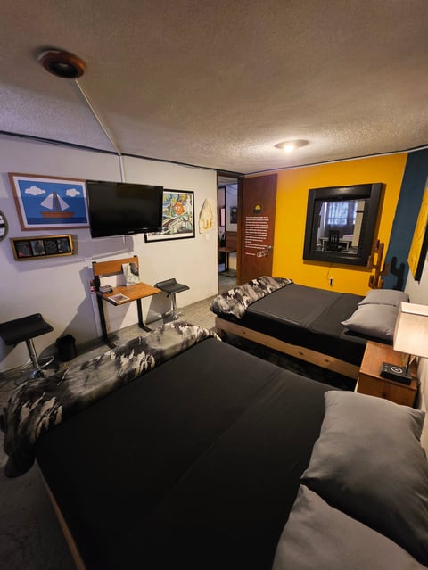 Modern bedroom with two black beds, wall-mounted TV, yellow accent wall, desk area, and warm lighting