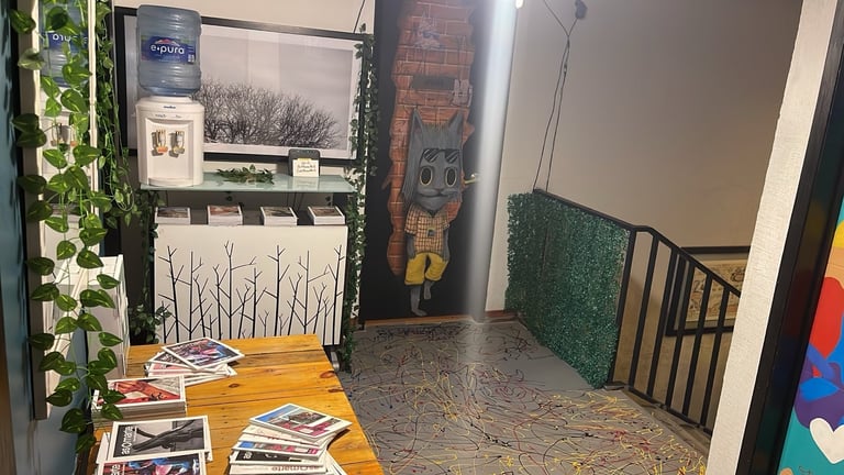 Art gallery or museum display space with framed artwork on walls, wooden table with printed materials, and decorative green plants and wall art
