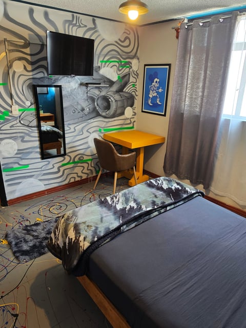 Modern bedroom with colorful abstract wall mural, yellow desk, wall-mounted TV, and bed with decorative bedding