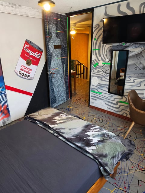 Bedroom interior with bed, pop art posters including Campbells soup can, open doorway with warm lighting, abstract wall art, and modern furnishings