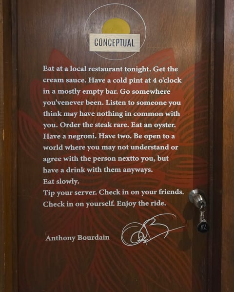 Wooden sign with Conceptual restaurant logo and Anthony Bourdain quote about dining, including life advice about eating, drinking, and human connection.