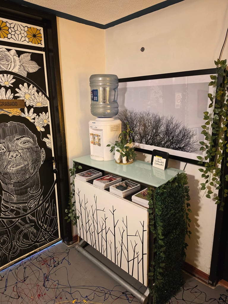 Modern office corner with water cooler, white cabinet with branch design, hanging plants, decorative wall art, and window with blinds