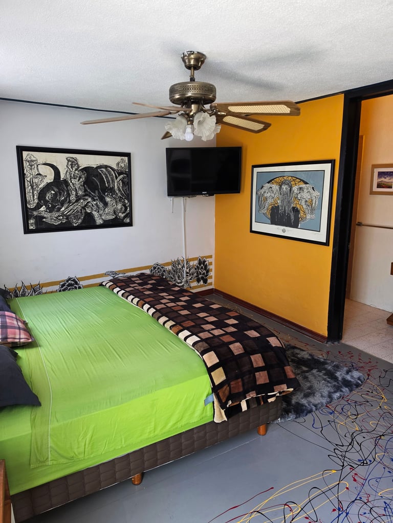 Bedroom with bright lime green bedding, bold yellow accent wall, ceiling fan with light fixture, black framed artwork, and wall-mounted TV.