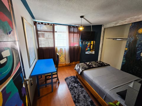 A colorful dorm room with two beds, blue desk, artwork on walls, window with dark curtains, and wooden flooring.